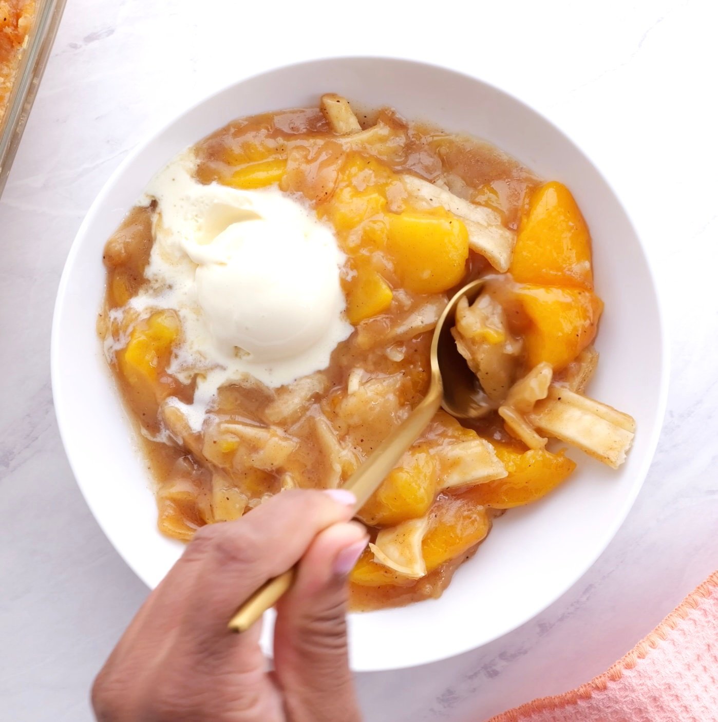 A white bowl of peach cobbler topped with ice cream with a hand dipping in.