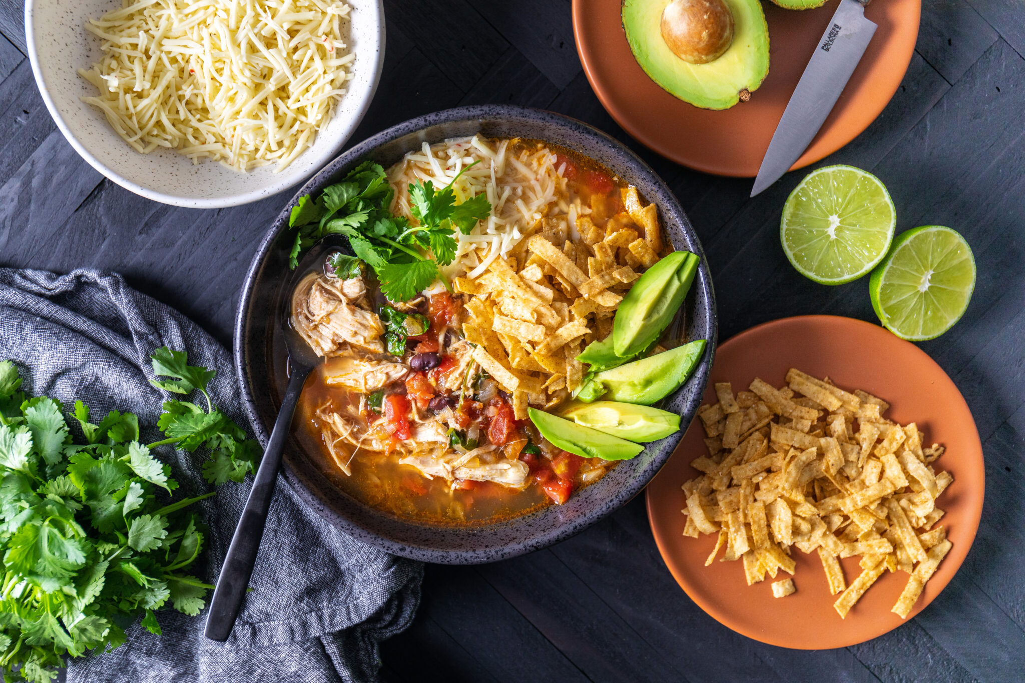 A bowl of chicken tortilla soup garnished with avocado slices, shredded cheese, and tortilla strips. Surrounding the bowl are lime halves on a plate, shredded cheese in a separate bowl, fresh cilantro, and a whole avocado with a knife on a cutting board.