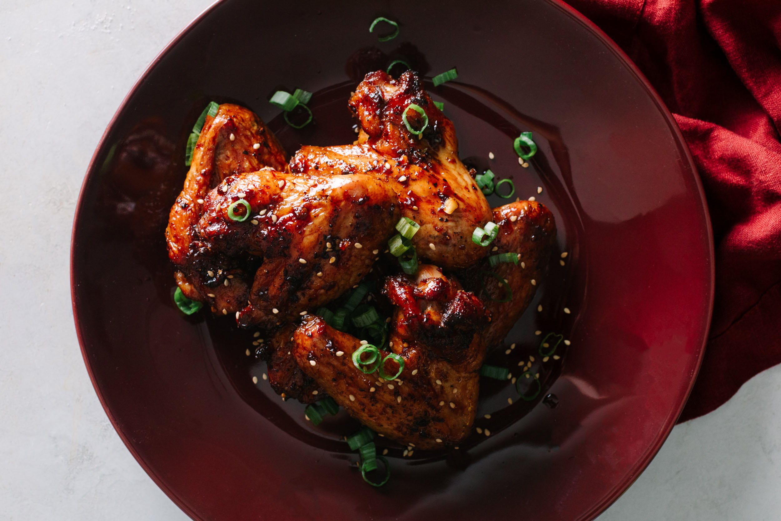 Asian chicken wings garnished with sesame seeds and green onions on a dark red plate.