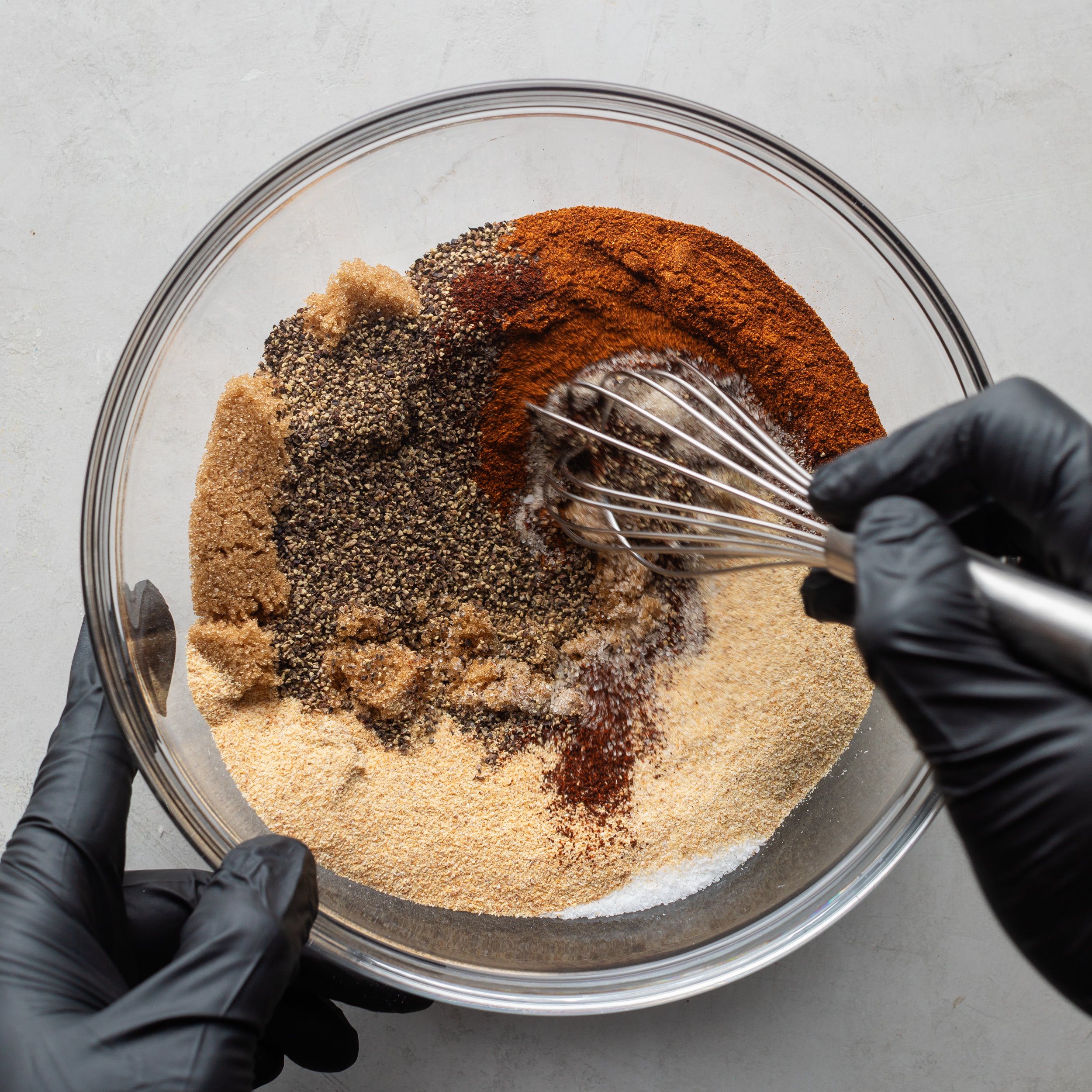 A hand wearing black latex gloves mixing with a whisk the BBQ seasoning ingredients in a large glass bowl.