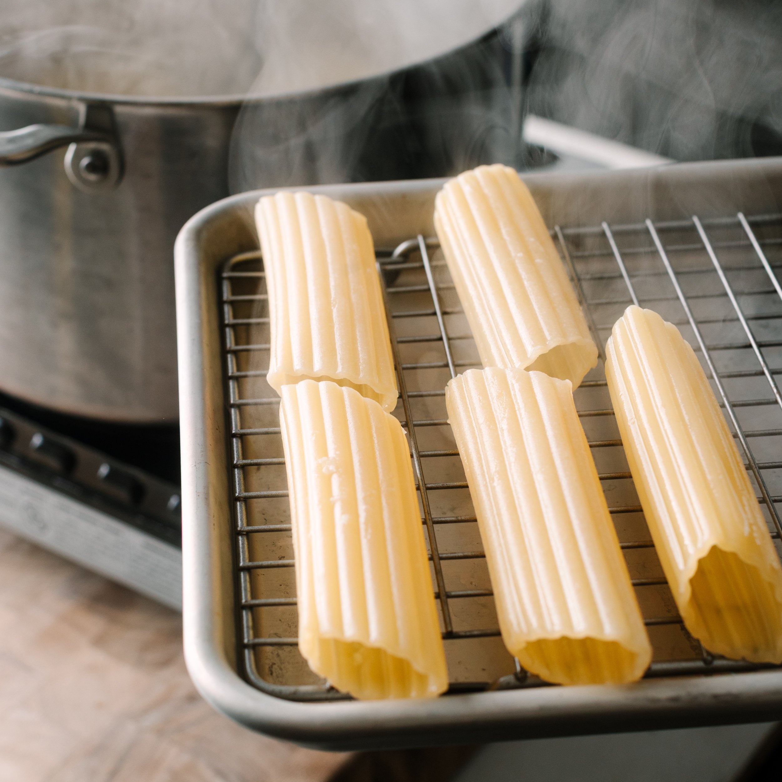 Cannelloni shells on a wire rack set over a sheet pan with a pot of boiling water in the background.