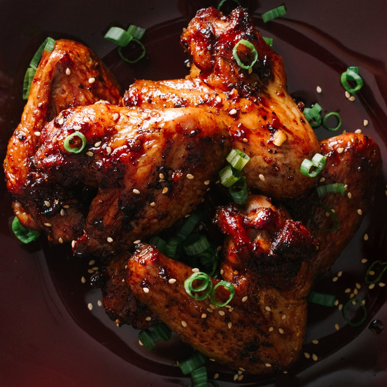 Asian chicken wings garnished with sliced green onions and toasted sesame seeds on a dark red plate.